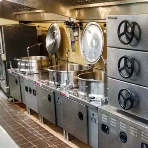 A row of stainless steam kettles under a hood along a kitchen wall